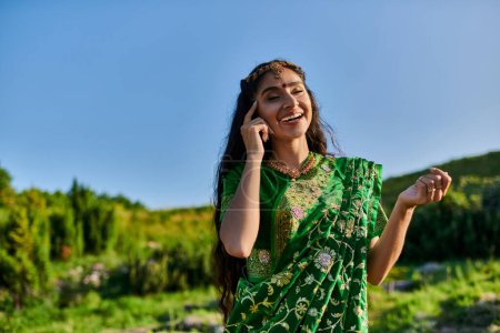 Téléchargez les photos : Joyeuse jeune femme indienne toucher le visage tout en posant en sari avec paysage sur fond - en image libre de droit