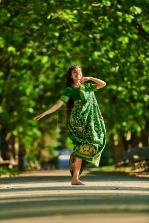 cheerful young indian woman in sari dancing on road with green trees on background