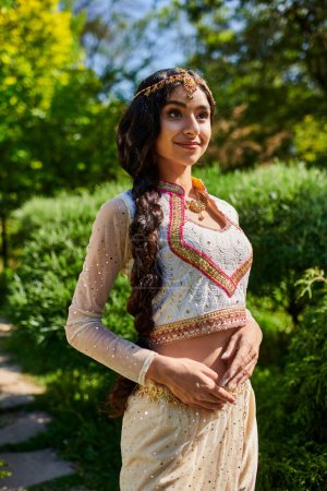 Téléchargez les photos : Élégant et jeune femme indienne en tenue traditionnelle à la mode debout et posant dans le parc - en image libre de droit
