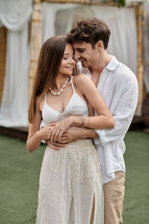 Foto de Guapo hombre abrazando tatuado novia en crop top y de pie juntos al aire libre, escapada de verano - Imagen libre de derechos