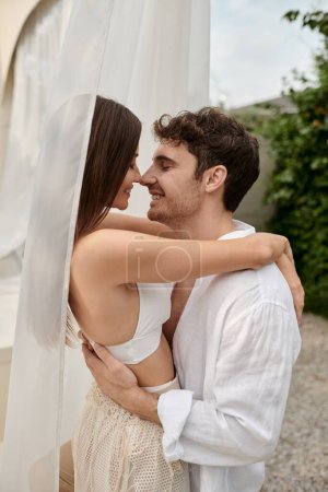 sexy Paar, glücklicher Mann umarmt schöne Frau, während sie in der Nähe von weißem Tüll des Pavillons am Strand steht