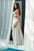 elegant woman with tattoo posing in bikini top and skirt near white tulle of beach house, serene mug #672029416