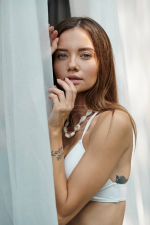 Téléchargez les photos : Portrait de femme avec tatouage et yeux bleus posant en haut de bikini près du tulle blanc de la maison de plage - en image libre de droit