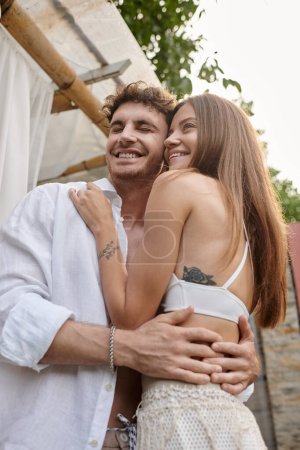 cheerful woman in white attire embracing with pleased man near beach pavilion during vacation