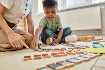 material montessori, chico afroamericano inteligente jugando juego educativo cerca del maestro, colorido