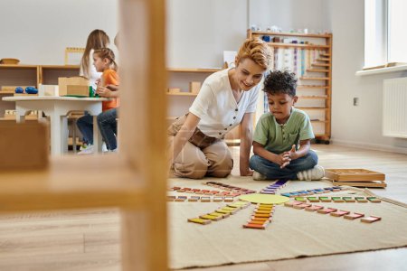 Montessori-Material, afrikanisch-amerikanischer Junge spielt pädagogisches Farbspiel in der Nähe fröhlicher Lehrer, Kinder