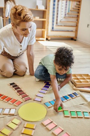 matériel montessori, garçon afro-américain intelligent jouant jeu de couleur éducatif près de femme heureuse