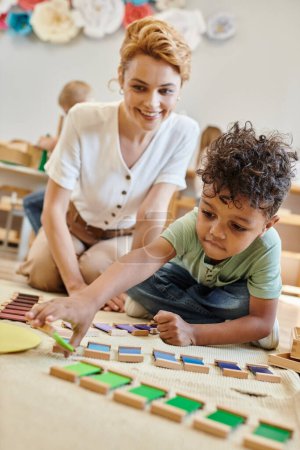 matériel montessori, garçon afro-américain intelligent jouant jeu de couleur éducatif près enseignant fier