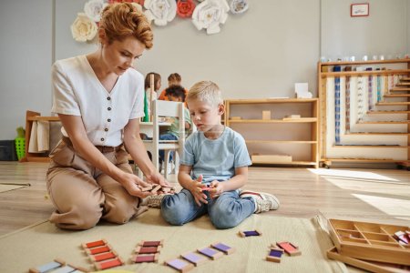 montessori school, female teacher sitting near blonde boy and showing wooden toys, educational game