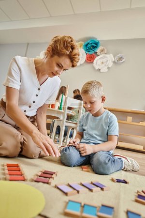montessori escuela, profesora sentada cerca de chico rubio jugando con juguetes de madera, juego educativo