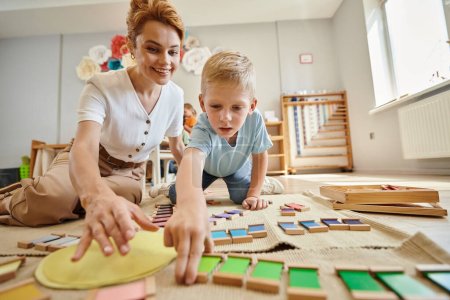 Montessori-Schule, blonder Junge spielt pädagogisches Spiel in der Nähe glücklicher Lehrer, Bewegung, Farbabstimmung