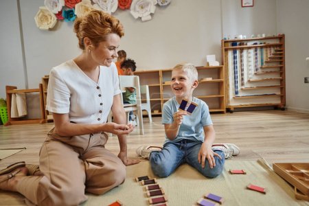 Montessori-Schule, glücklicher Junge beim Farbabgleich in der Nähe einer Lehrerin, sitzt auf dem Boden