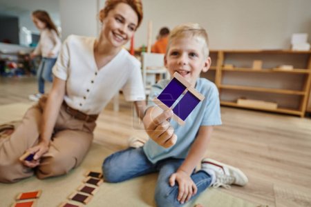 Montessori-Schule, fröhlicher Junge beim Farbenspiel in der Nähe einer Lehrerin, der auf dem Boden sitzt