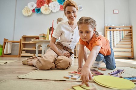 Montessori-Material, Mädchen spielt Farbabgleich-Spiel in der Nähe freudiger Lehrerin, die auf dem Boden sitzt