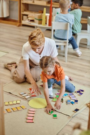 Montessori-Material, Mädchen spielen farblich abgestimmtes Spiel in der Nähe freudiger Lehrerin, diverse Jungen