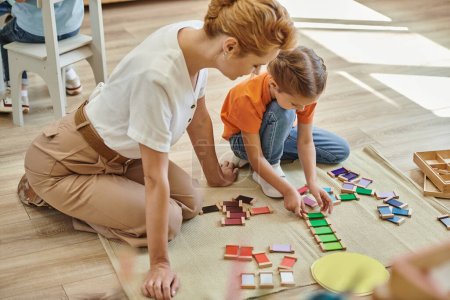 Montessori-Konzept, Mädchen spielt Farbabgleich-Spiel in der Nähe freudiger Lehrerin, die auf dem Boden sitzt