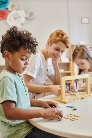 afrikanisch-amerikanischer Junge, der Kacheln zählt und verwendet, Montessori-Konzept, Lehrer und Mädchen spielen Spiel
