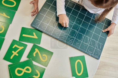 vista superior de la niña escribiendo número cero en pizarra, aprendiendo a contar en la escuela Montessori