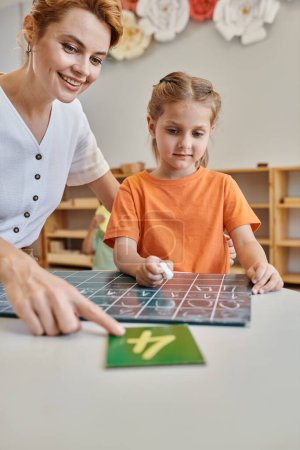 Glückliche Lehrerin zeigt auf Zahl neben intelligentem Mädchen in Montessori-Schule, Zählen, Schreiben, Kreidetafel