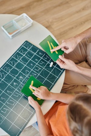 girl and teacher pointing at number near chalkboard, learning through play, counting, cropped