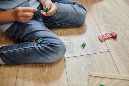 vista recortada de niño sentado en jeans en el suelo, jugando con material de cuentas Montessori, juego