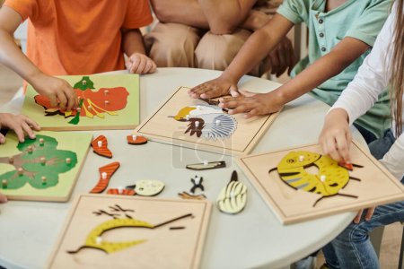 abgeschnittene Ansicht von interrassischen Kindern, die mit didaktischen Materialien in der Nähe des Lehrers in der Montessori-Schule spielen