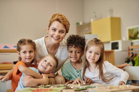 Lächelnder Lehrer und multiethnische Kinder blicken während des Unterrichts in der Montessori-Schule in die Kamera