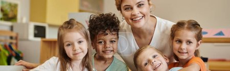 profesor positivo y niños interracial mirando a la cámara en la escuela montessori, pancarta