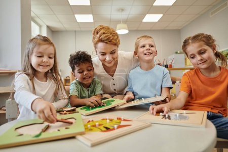fröhlicher Lehrer, der in der Montessori-Klasse neben multiethnischen Kindern sitzt, die mit didaktischem Material spielen