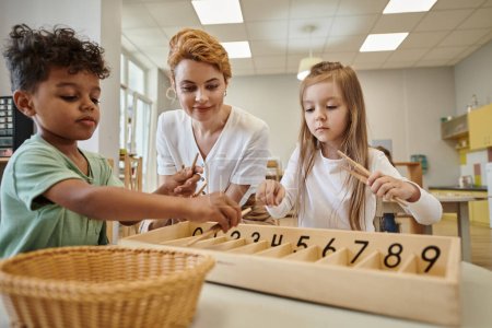 multiethnische Kinder spielen in der Montessori-Schule neben lächelndem Lehrer mit Holzstäben