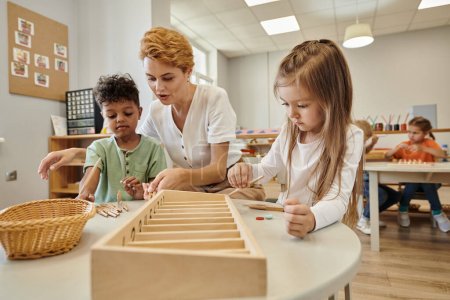 teacher talking to multiethnic kids while playing with wooden sticks in montessori school