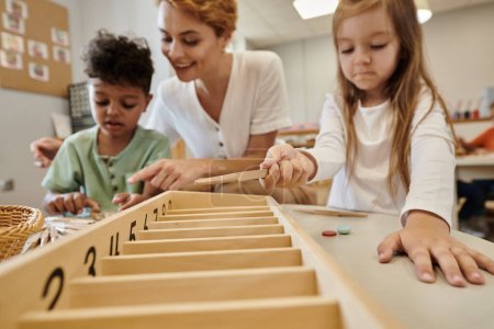 niños confusos multiétnicos jugando con palos de madera cerca del profesor en la escuela montessori
