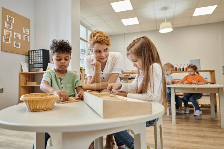 teacher sitting near multiethnic kids playing with wooden sticks during lesson in montessori school