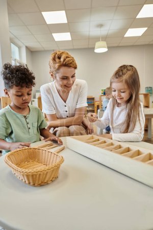 positive teacher looking at multiethnic kids playing with wooden sticks in montessori school