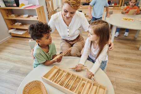 high angle view of smiling teacher hugging multiethnic kids playing with sticks in montessori school