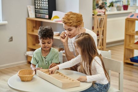 smiling multiethnic kids playing with wooden sticks while teacher talking in montessori school