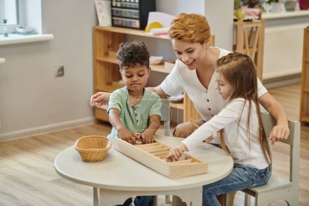 glücklicher Lehrer im Gespräch mit interrassischen Kindern, die in der Montessori-Klasse mit didaktischen Materialien aus Holz spielen