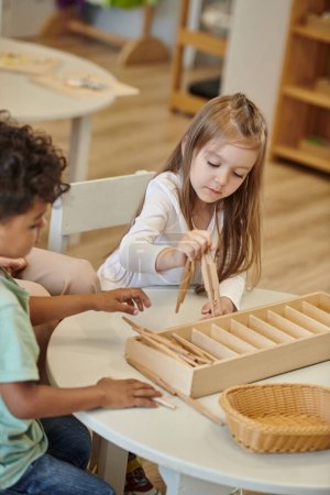Foto de Niños interracial jugando con palos de madera en la mesa durante la lección en la escuela montessori - Imagen libre de derechos
