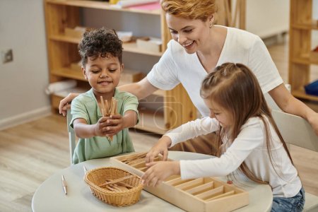 niños multiétnicos positivos jugando con materiales didácticos de madera cerca del profesor en la escuela montessori