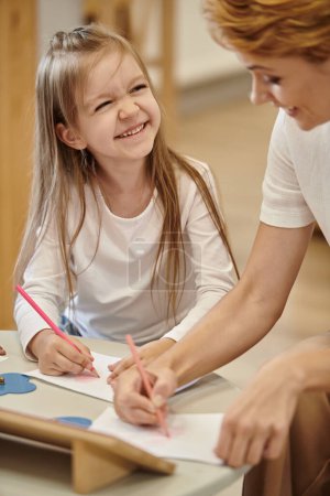 alegre chica pintura y mirando borrosa profesor durante lección en montessori escuela