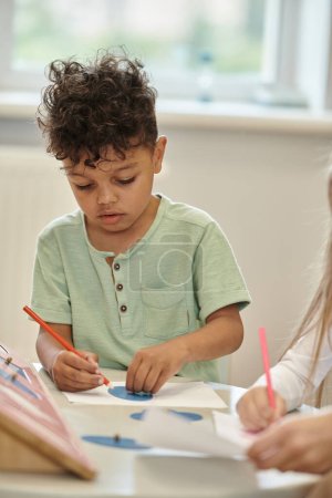 Photo for African american child paining with pencil near friend and blurred teacher in montessori class - Royalty Free Image