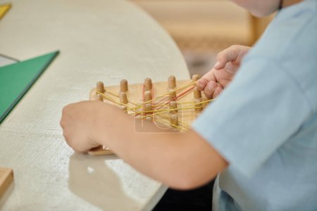 Ausgeschnittene Ansicht eines Jungen, der mit Gummibändern und Holzstäben auf einem Tisch in der Montessori-Schule spielt