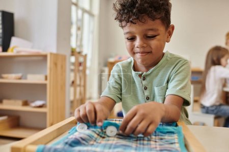 niño afroamericano sonriente jugando con tela y botones en la escuela montessori