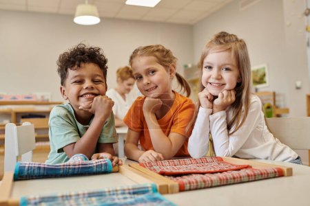 Foto de Niños alegres y multiétnicos mirando la cámara cerca de la mesa en la clase de la escuela montessori - Imagen libre de derechos