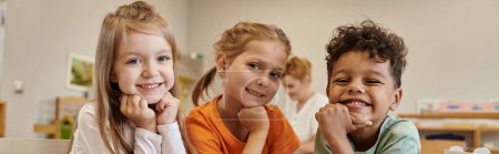 niños sonrientes e interracial mirando a la cámara en clase borrosa en la escuela montessori, pancarta