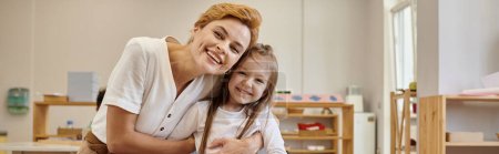 positive teacher and pupil looking at camera together in class in montessori school, banner