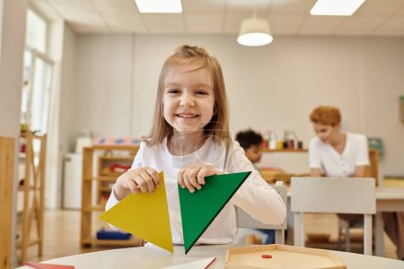 niño alegre sosteniendo triángulos y mirando a la cámara en clase en la escuela montessori