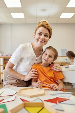 positiver Lehrer umarmt Mädchen und schaut in die Kamera in der Klasse in der Montessori-Schule