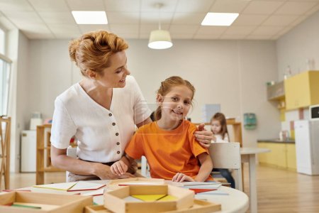 Téléchargez les photos : Professeur souriant étreignant fille près du matériel didactique dans la classe de l'école montessori - en image libre de droit