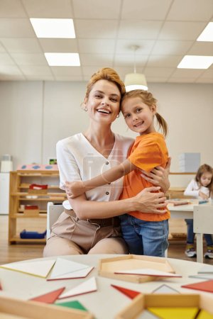 positive pupil hugging teacher and looking at camera in class of montessori school